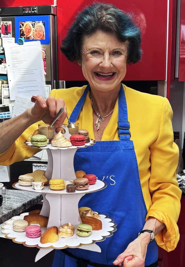 Rosy smiling, holding a display of petite fours, wearing a yellow jacket.