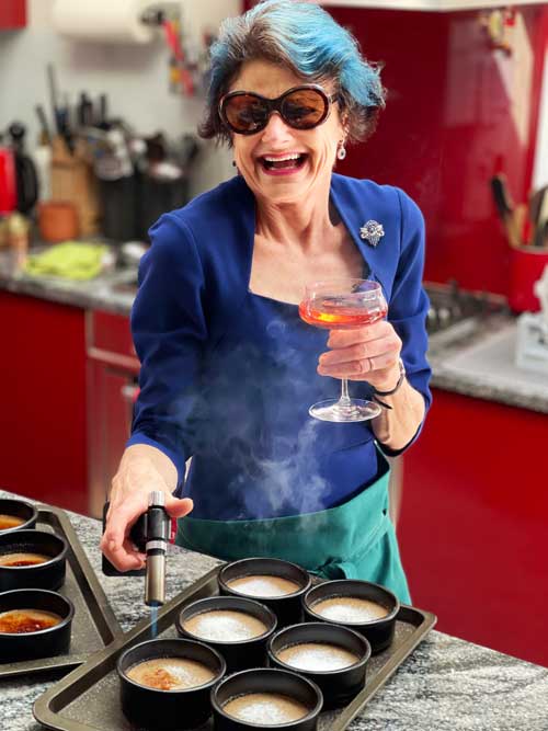 Rosy, wearing sunglasses and with a cocktail in her hand, laughing while she operates a blowtorch on dessert bowls.