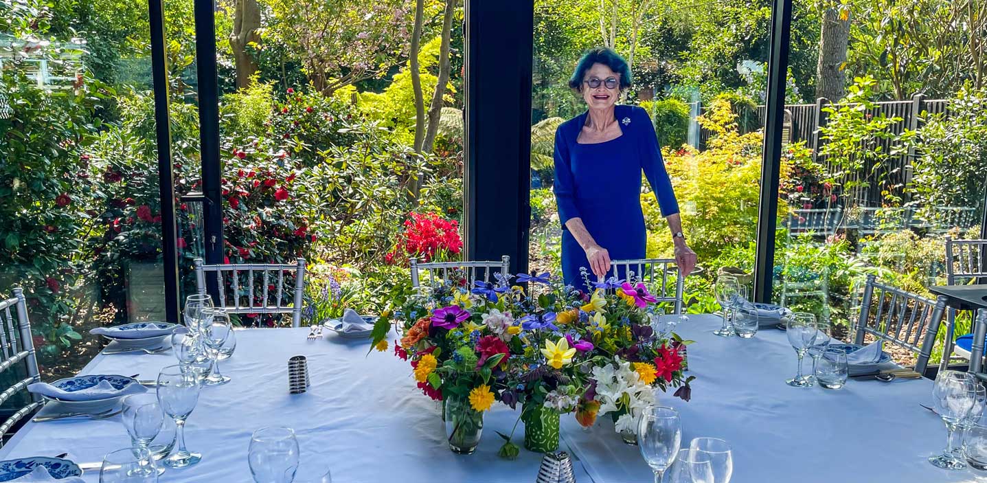 Rosy smiling in the pavilion, standing behind a beautifully set table with gorgeous, multi-coloured flower vases on it.
