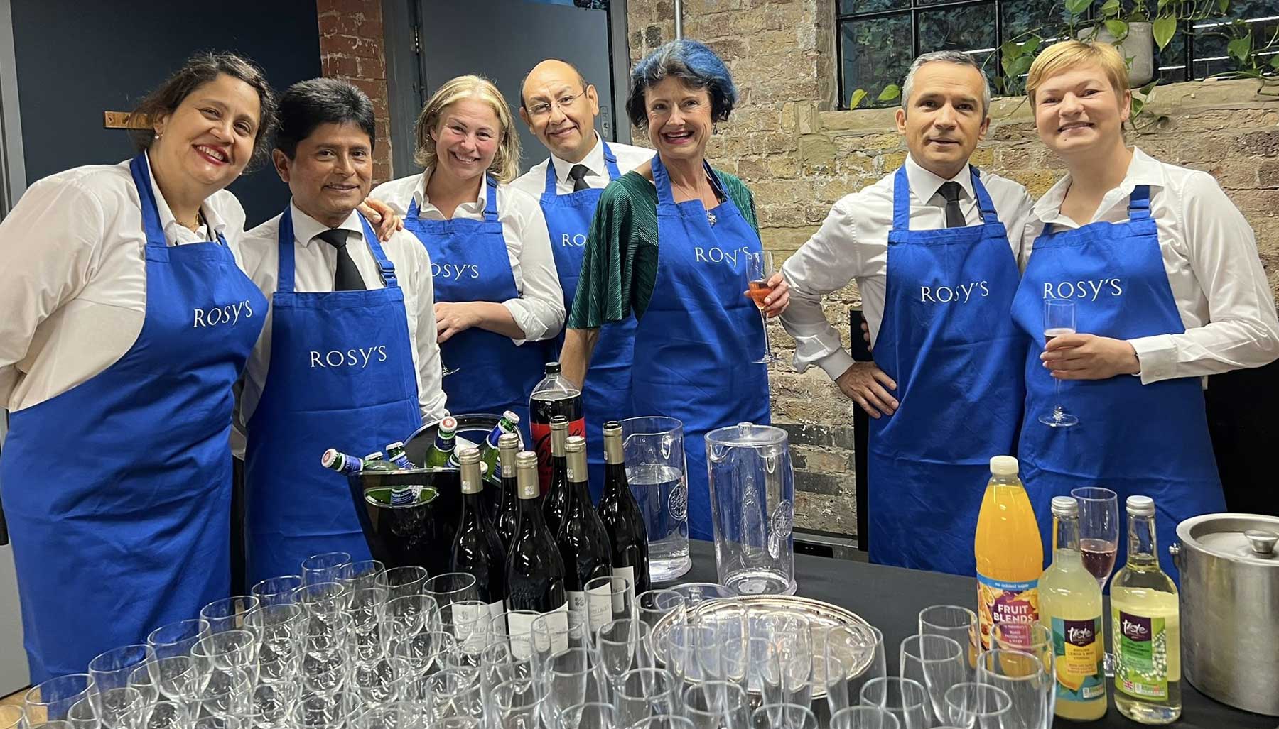 Rosy and her team, happily smiling behind a table laid out with drinks and glasses.