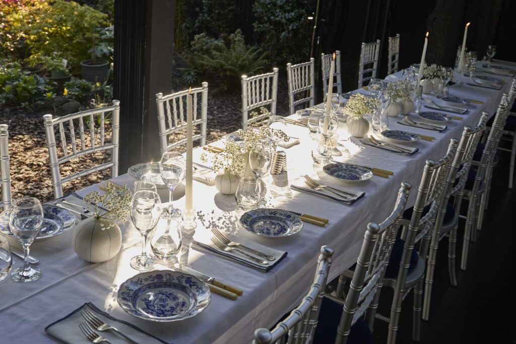 Diagonal view of a beautifully set table in the pavilion in the daytime, with the garden in the background and the sun shining on it.