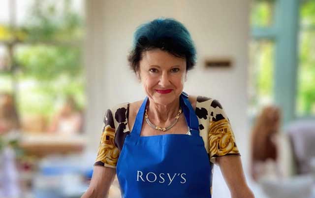 Rosy looking at the camera with a semi-smile on her face, wearing a blue apron with her logo on it.